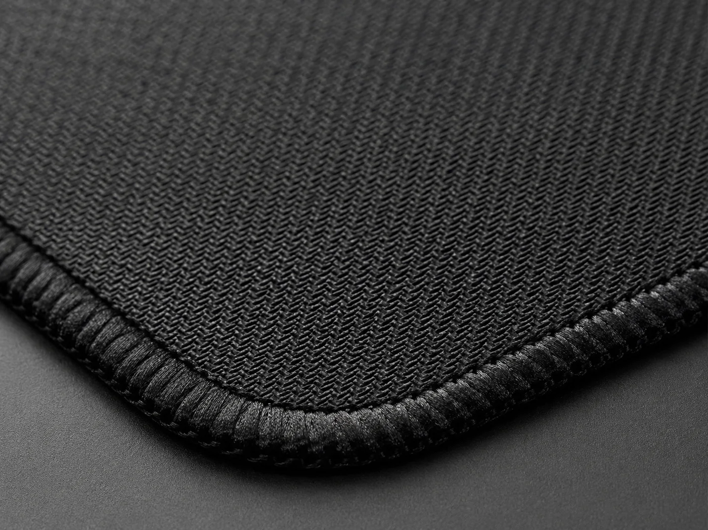 Macro close-up of a black desk mat corner showing the fine fabric weave and the tight stitched edge running around the rounded corner
